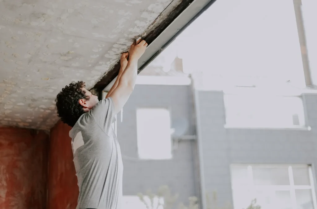 A person in a gray T-shirt reaches up, adjusting a ceiling panel in a softly lit room. The scene conveys focus and DIY effort with a serene atmosphere.