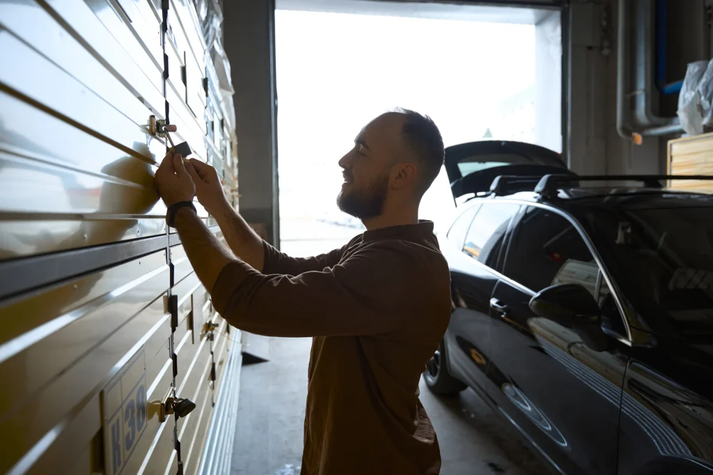 A man in a brown shirt locks a large storage unit with a padlock, smiling. A black car with an open hatchback is parked nearby in a dimly lit garage.