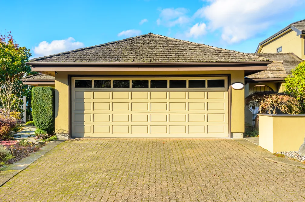 A suburban home with a two-car garage features a beige garage door with window panels. Surrounding greenery and clear blue sky create a serene atmosphere.