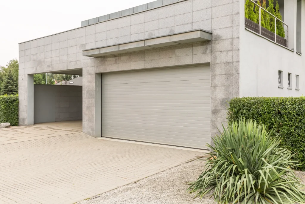 Modern house exterior featuring a gray stone facade with a closed garage door. Neatly trimmed bushes and plants line the driveway, creating a tidy and minimalist appearance.