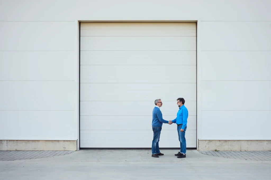 Two men shake hands in front of a large, closed white garage door. Both wear blue shirts and jeans, conveying a sense of casual cooperation.