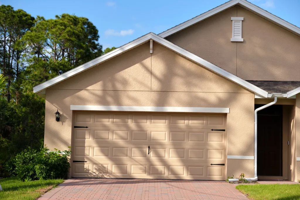 The image shows a beige house with a two-car garage and beige panels. Shadows of trees create patterns on the wall. There is a red brick driveway and greenery nearby.