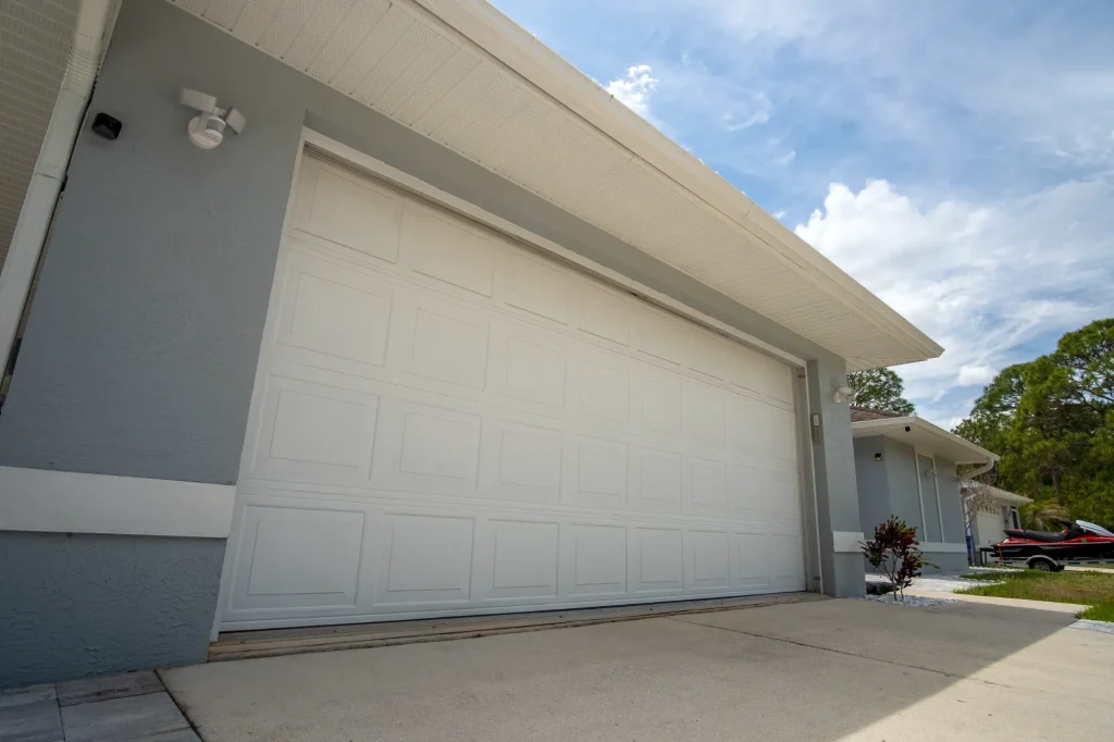 Angled view of a modern, single-car garage with a white door on a light blue house under a partly cloudy sky. The driveway is empty, conveying tranquility.