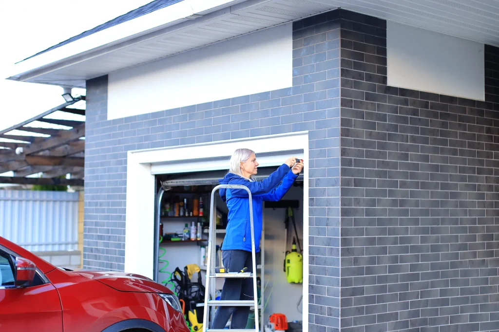A person in a blue jacket on a ladder uses a screwdriver to repair a garage door, with a parked red car in the foreground. The mood is focused and industrious.